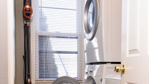 A compact laundry room with a front-loading washer and dryer, a window with blinds, a vacuum cleaner, and a black laundry basket. - Geauga Maple Cottage (Chardon)