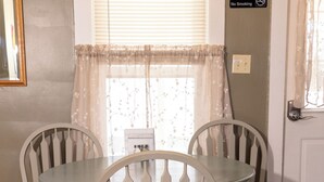 A dining area with a table and four chairs, a window with curtains, a 'No Smoking' sign, and a door with a doorknob. - Geauga Maple Cottage (Chardon)