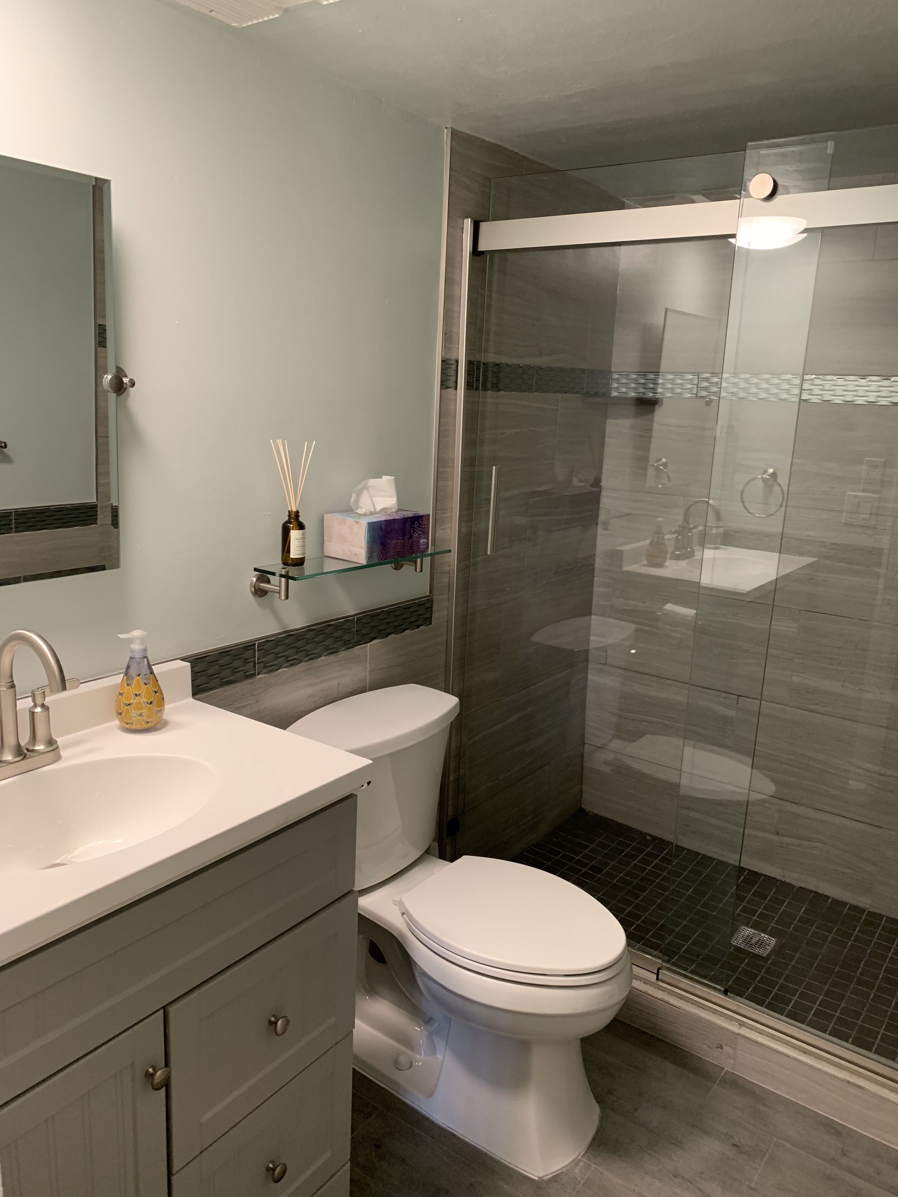 Une salle de bain moderne avec une douche à parois de verre, des toilettes blanches et un meuble-lavabo doté d’un miroir et d’un distributeur de savon.