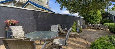 Un área de patio al aire libre con una mesa redonda y sillas, una pared negra y un árbol que da sombra.