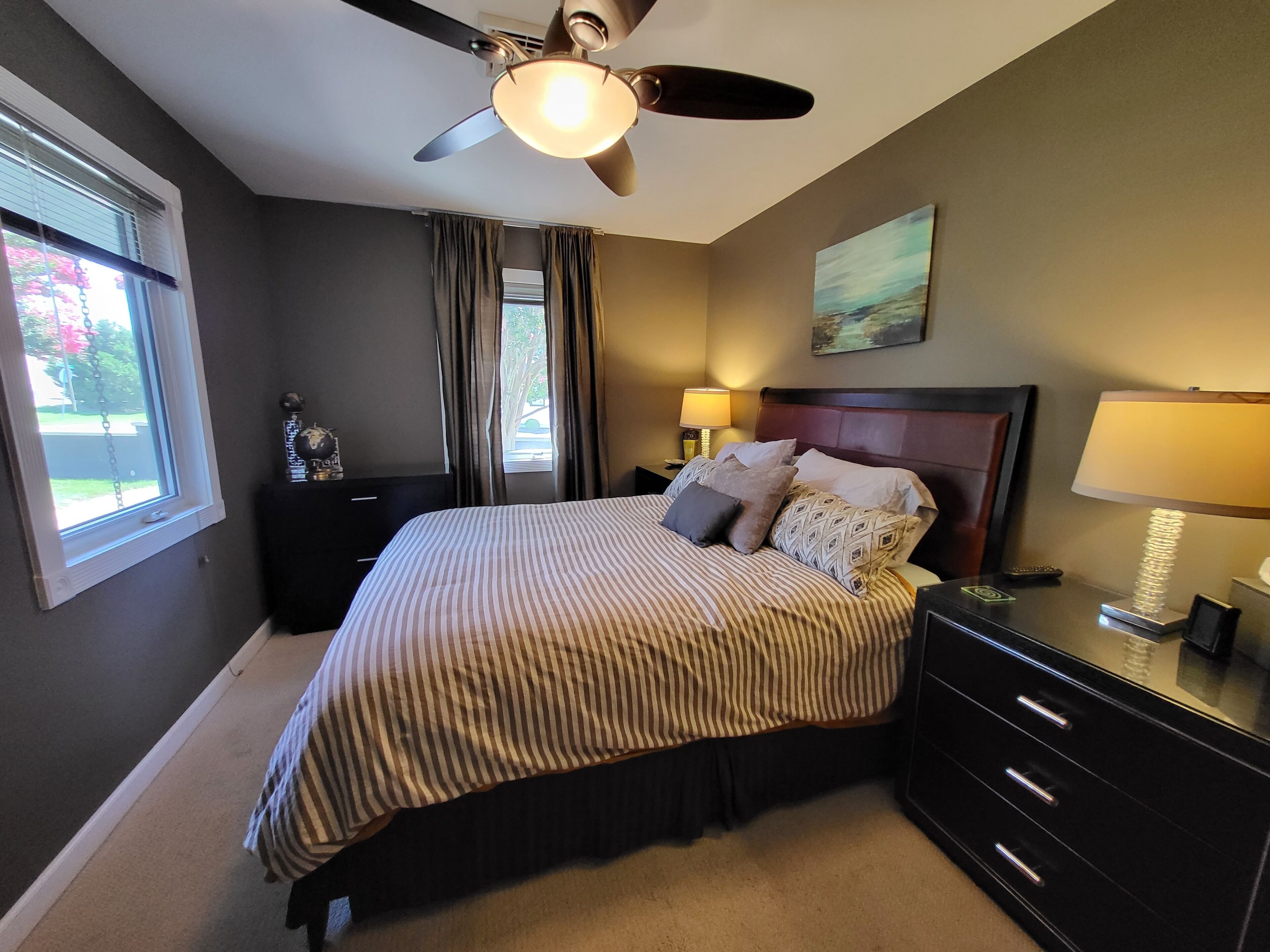 A bedroom with a bed, a ceiling fan, a nightstand, a lamp, a dresser, and a window with curtains.