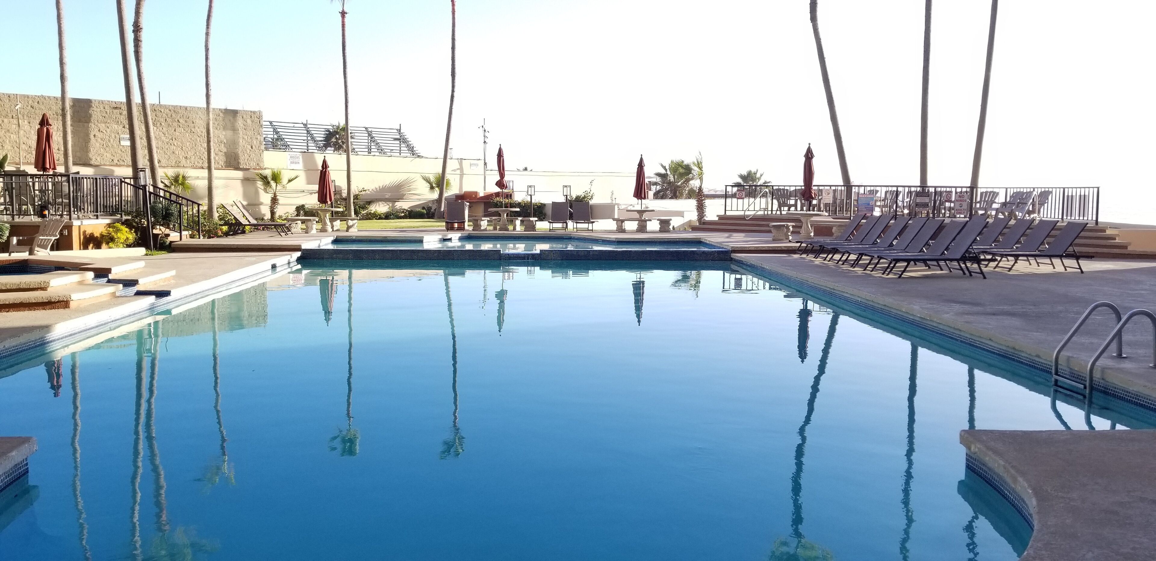 Una piscina cristallina con lettini e ombrelloni, circondata da palme e un edificio con una terrazza.