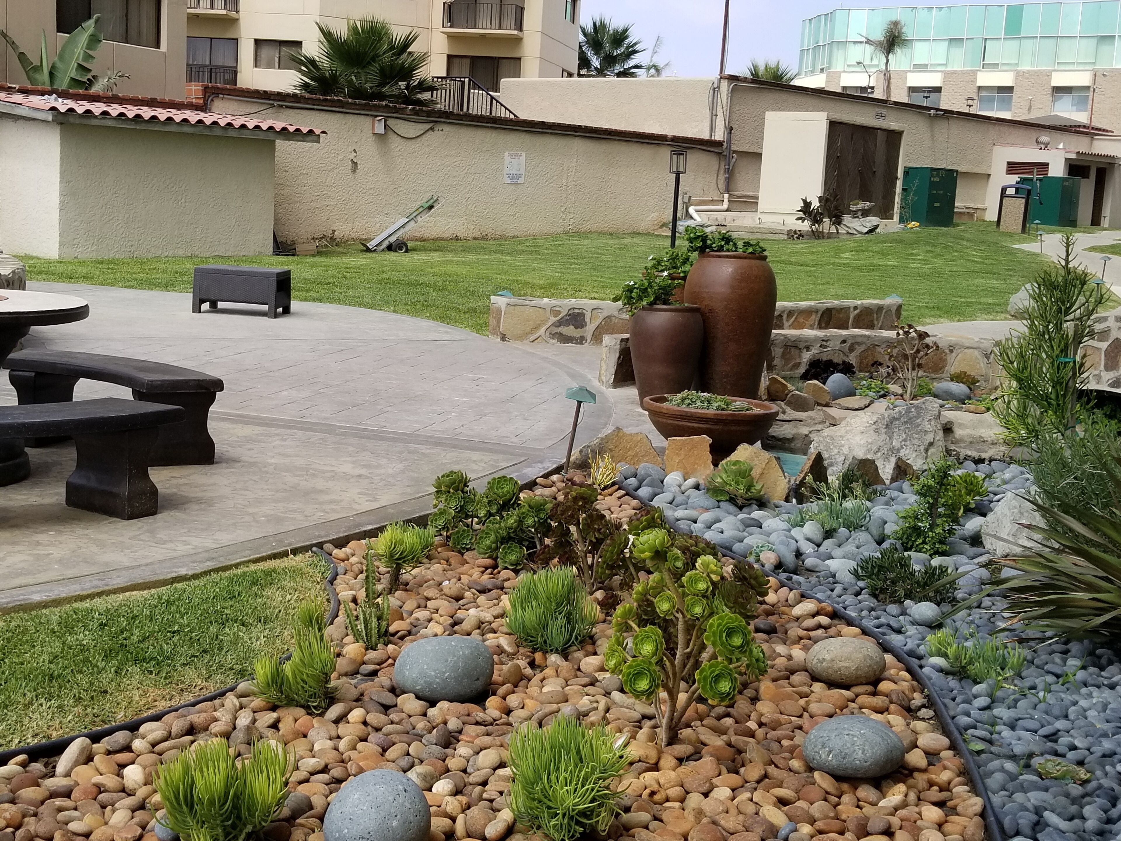 A patio area with a round table and benches, surrounded by a garden with various plants and rocks.