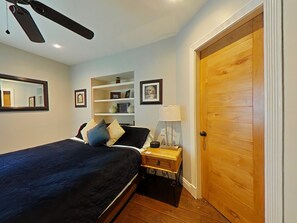 Quarto com cama, mesa de cabeceira, abajur, porta de madeira e prateleira com quadros emoldurados.