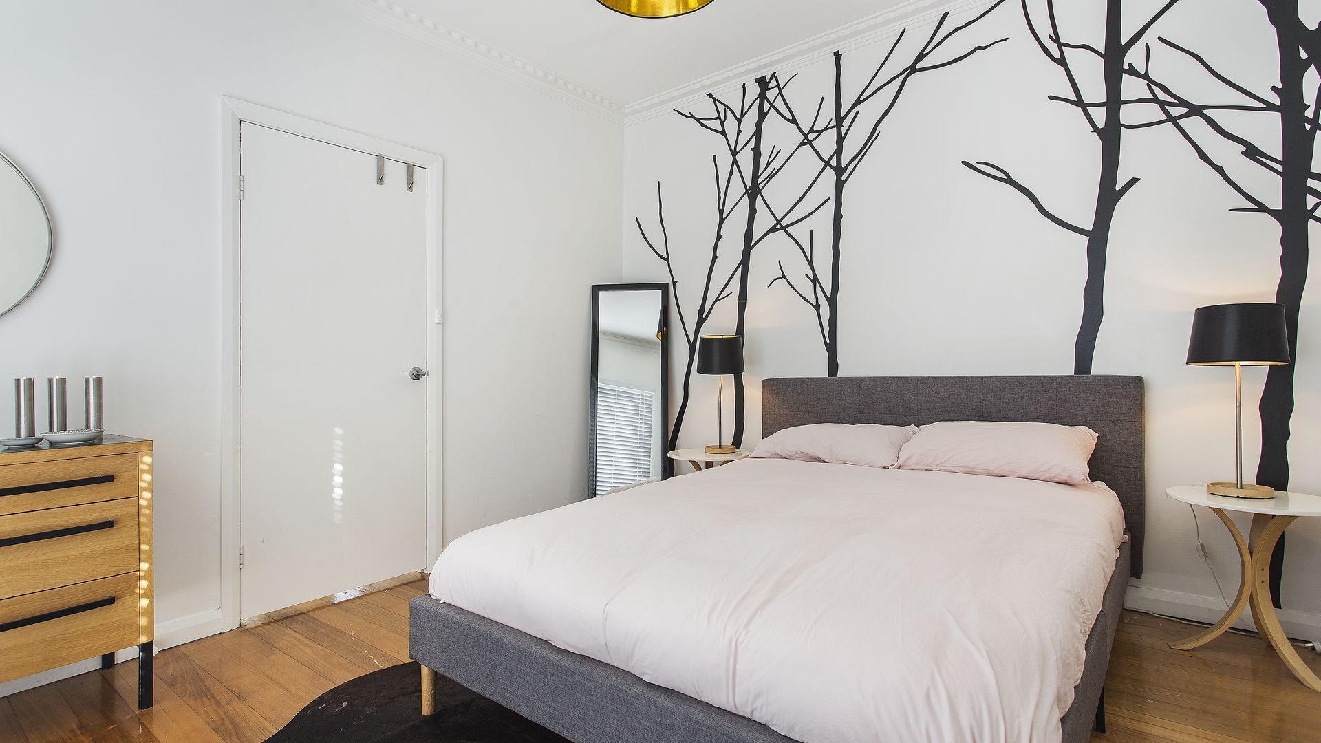 A bedroom with a bed, bedside tables, a dresser, a mirror, and a wall with a tree design.