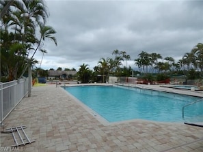 Une piscine dotée d’une terrasse carrelée, entourée de palmiers et surplombée par un ciel nuageux.