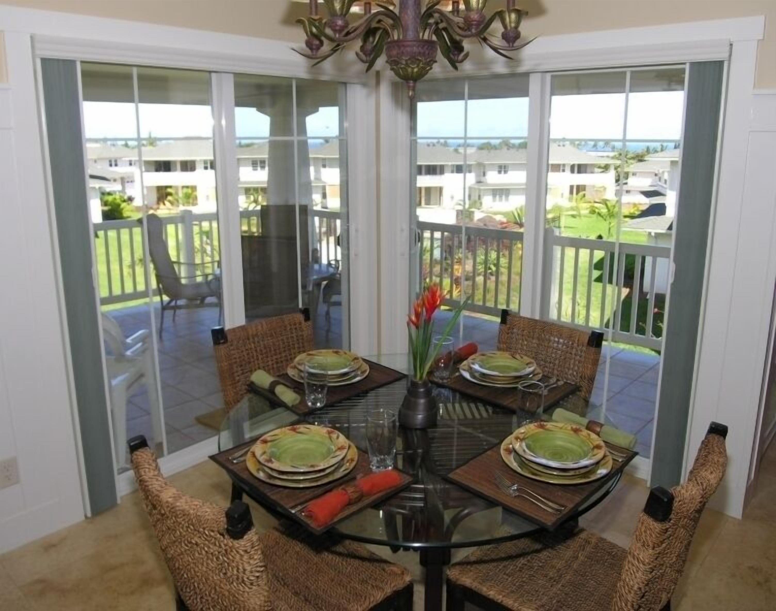 Un coin repas avec une table en verre entourée de chaises en osier, un lustre et une vue sur un paysage verdoyant, visible à travers de grandes portes-fenêtres coulissantes.