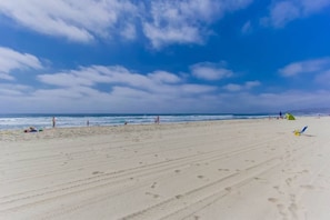 Una amplia playa de arena con huellas que conducen al agua, un cielo despejado y algunas personas a lo lejos.