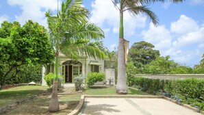 Una casa con entrada pavimentada, un pequeño jardín con palmeras y césped.