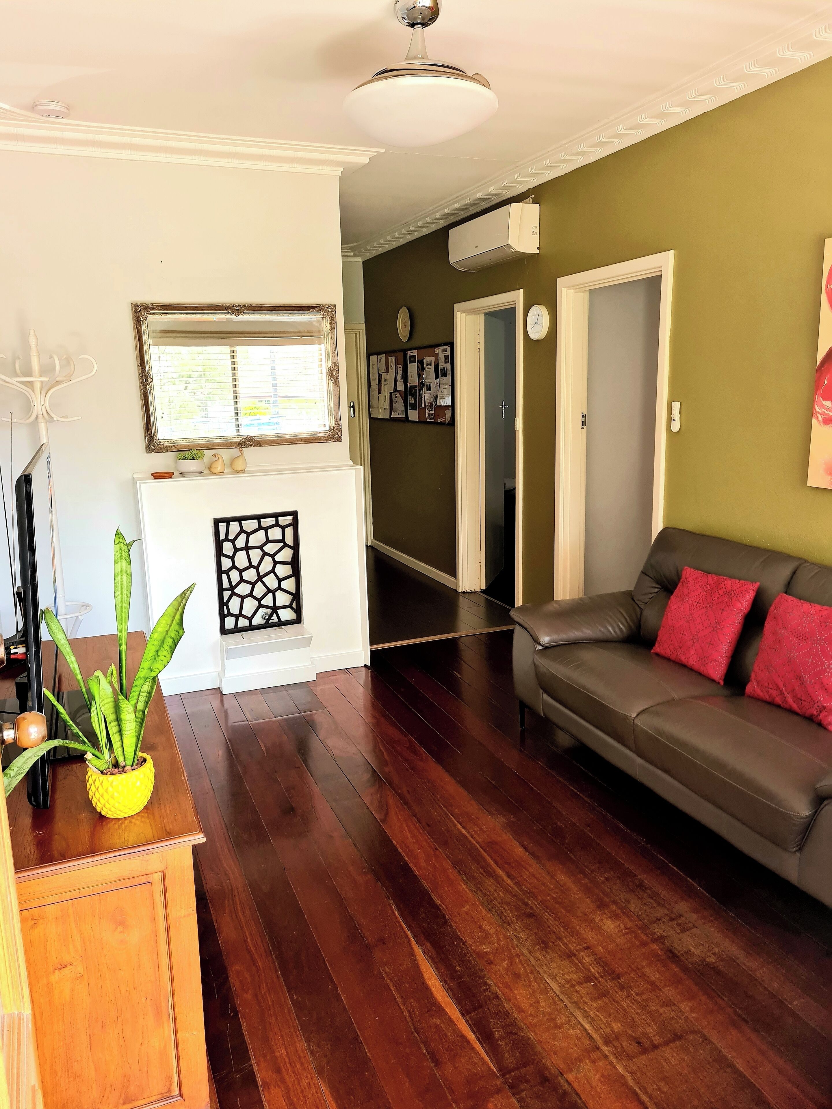 A living room with a dark wooden floor, a grey sofa with red cushions, a white fireplace, a ceiling fan, and a green wall.