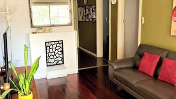 A living room with a dark wooden floor, a grey sofa with red cushions, a white fireplace, a ceiling fan, and a green wall.