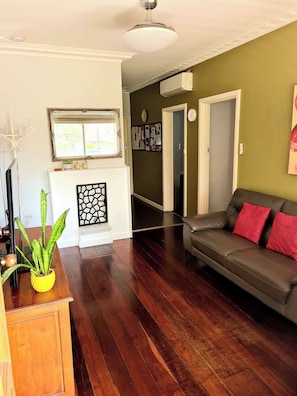 A living room with a dark wooden floor, a grey sofa with red cushions, a white fireplace, a ceiling fan, and a green wall.
