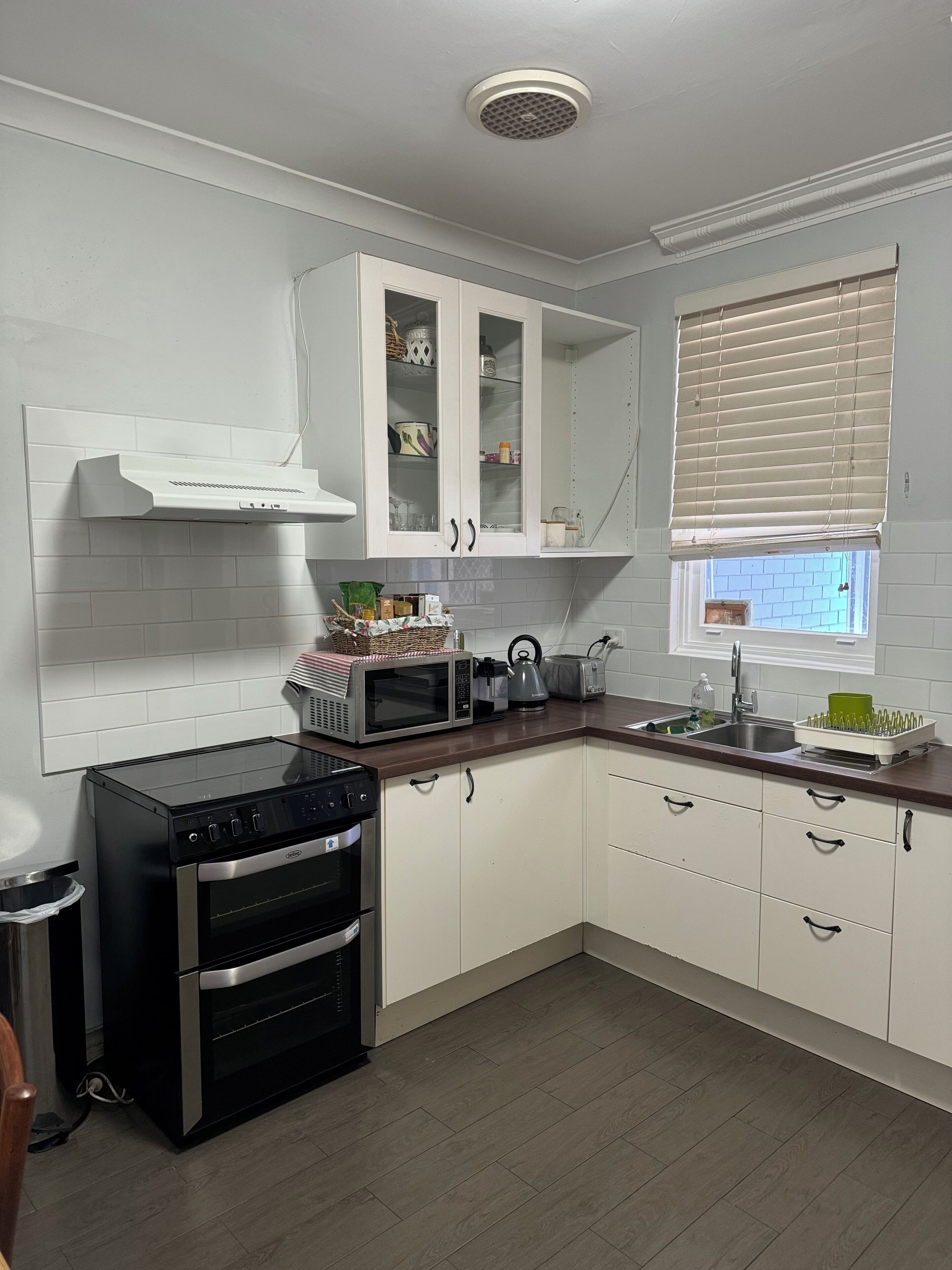 A modern kitchen with white cabinets, a built-in oven, a microwave, and a window with blinds.