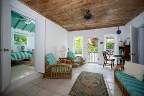 A room with a bed, two wicker chairs, a table, a lamp, and a view of greenery outside. - Poponi Villa on pink sand Beachfront Estate (North Palmetto Point)