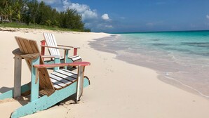 Una playa con dos sillas de madera tipo Adirondack y una mesa, cielo azul despejado y mar tranquilo.