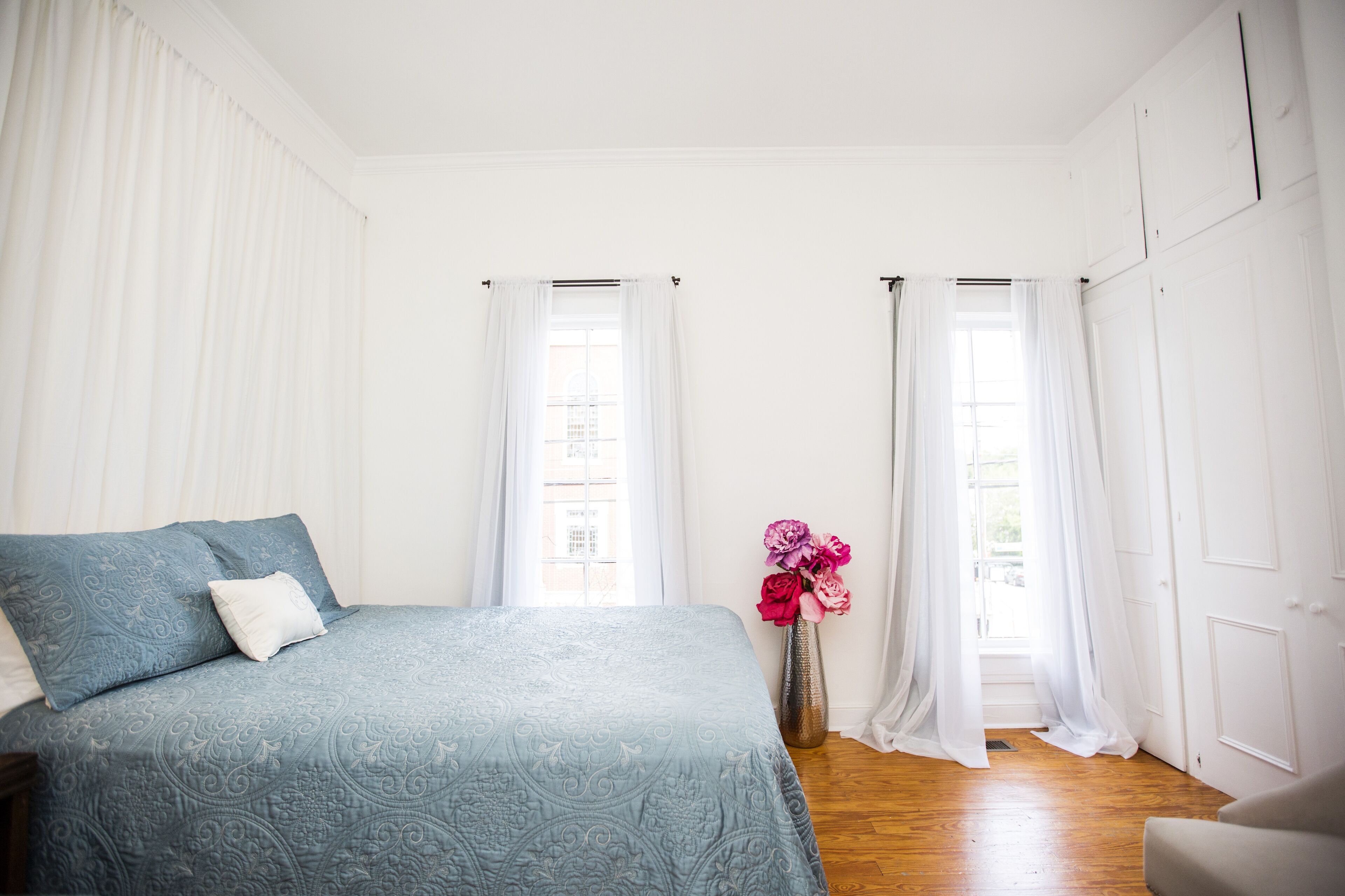 A bedroom with a bed, a vase with flowers, and two windows with curtains.