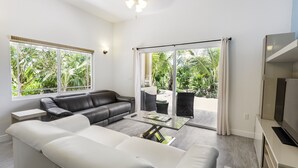 A modern living room with a white sofa, a black leather couch, a glass coffee table, a flat-screen TV, and a sliding glass door leading to a balcony with a view of greenery. - Sea La Vie - Beachside 1 Bedroom Unit #5 (Providenciales)