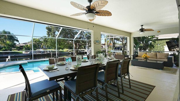 Un espace repas avec vue sur la piscine, de grandes fenêtres, des ventilateurs de plafond et des sièges d’extérieur.