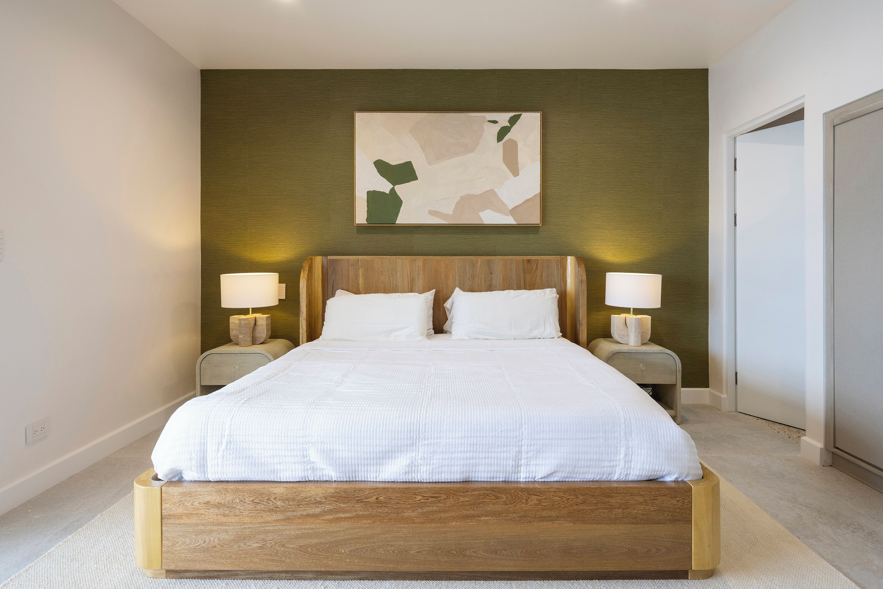 Uma cama grande com estrutura de madeira e roupa de cama branca, ladeada por duas mesas de cabeceira com luminárias, em um quarto com uma parede verde e uma obra de arte abstrata sobre a cama.