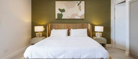Uma cama grande com estrutura de madeira e roupa de cama branca, ladeada por duas mesas de cabeceira com luminárias, em um quarto com uma parede verde e uma obra de arte abstrata sobre a cama.