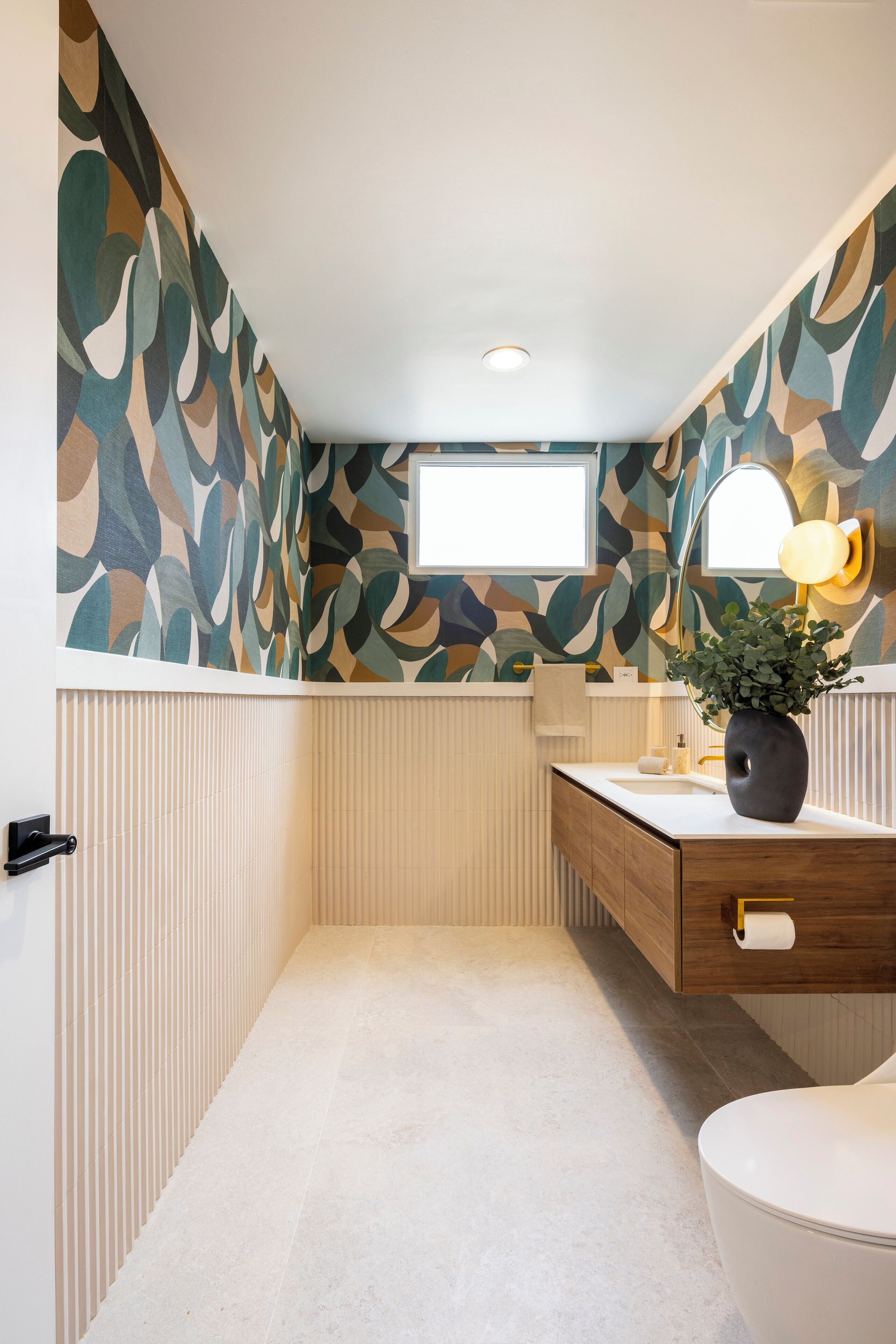 A bathroom with a patterned wall, a wooden vanity, a white toilet, and a mirror with a light.
