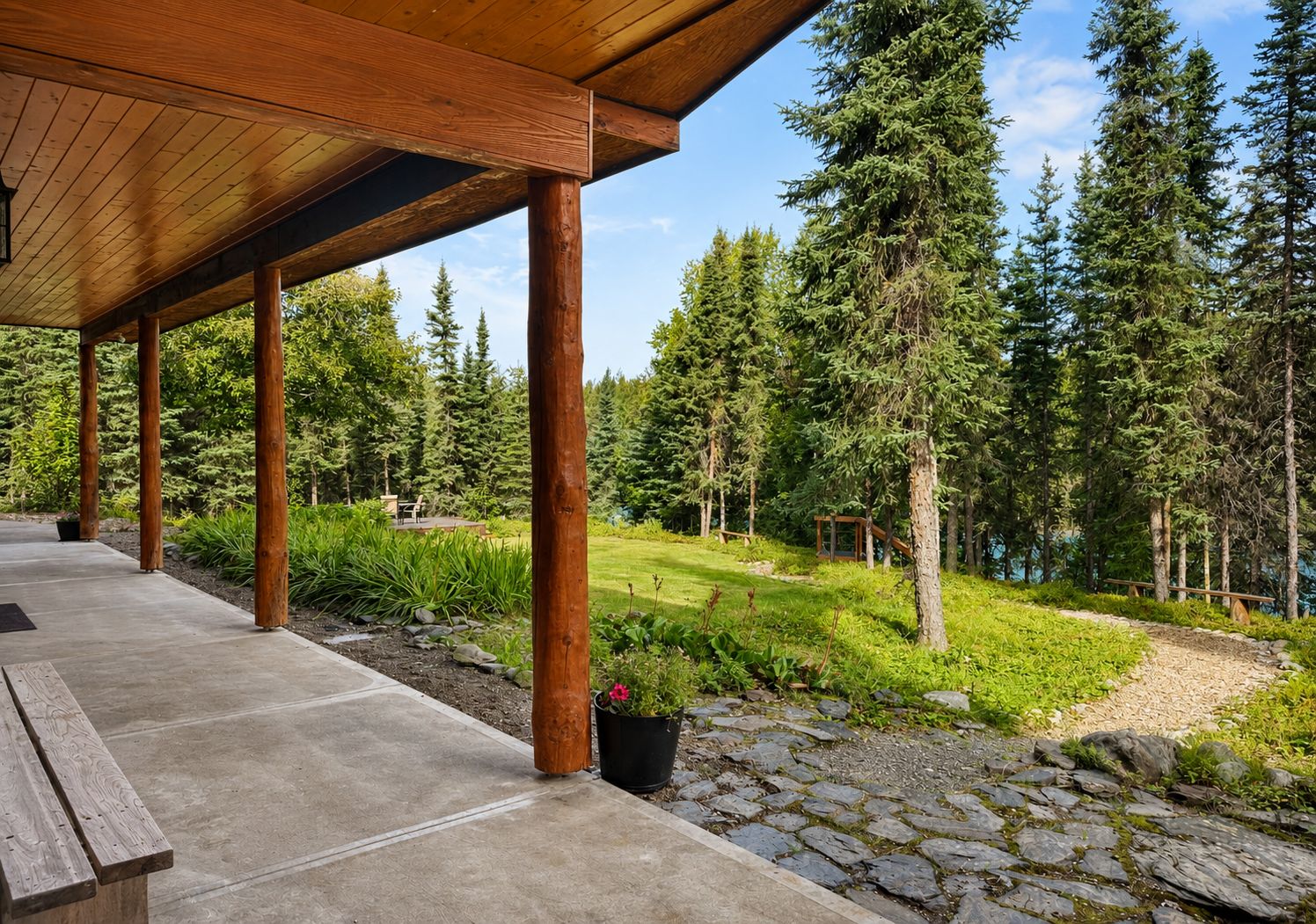 A covered outdoor area with a concrete floor, wooden pillars, and a stone pathway leading to a natural landscape with trees and a body of water.