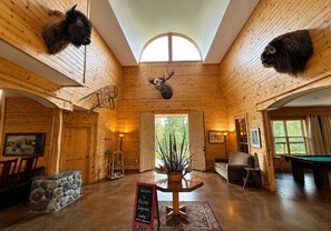 A lodge interior with wooden walls, a central fireplace, a billiards table, and mounted animal heads.