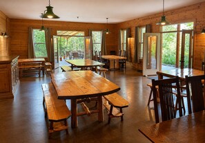 A rustic wooden interior with multiple dining tables and benches, large windows with curtains, and hanging pendant lights.