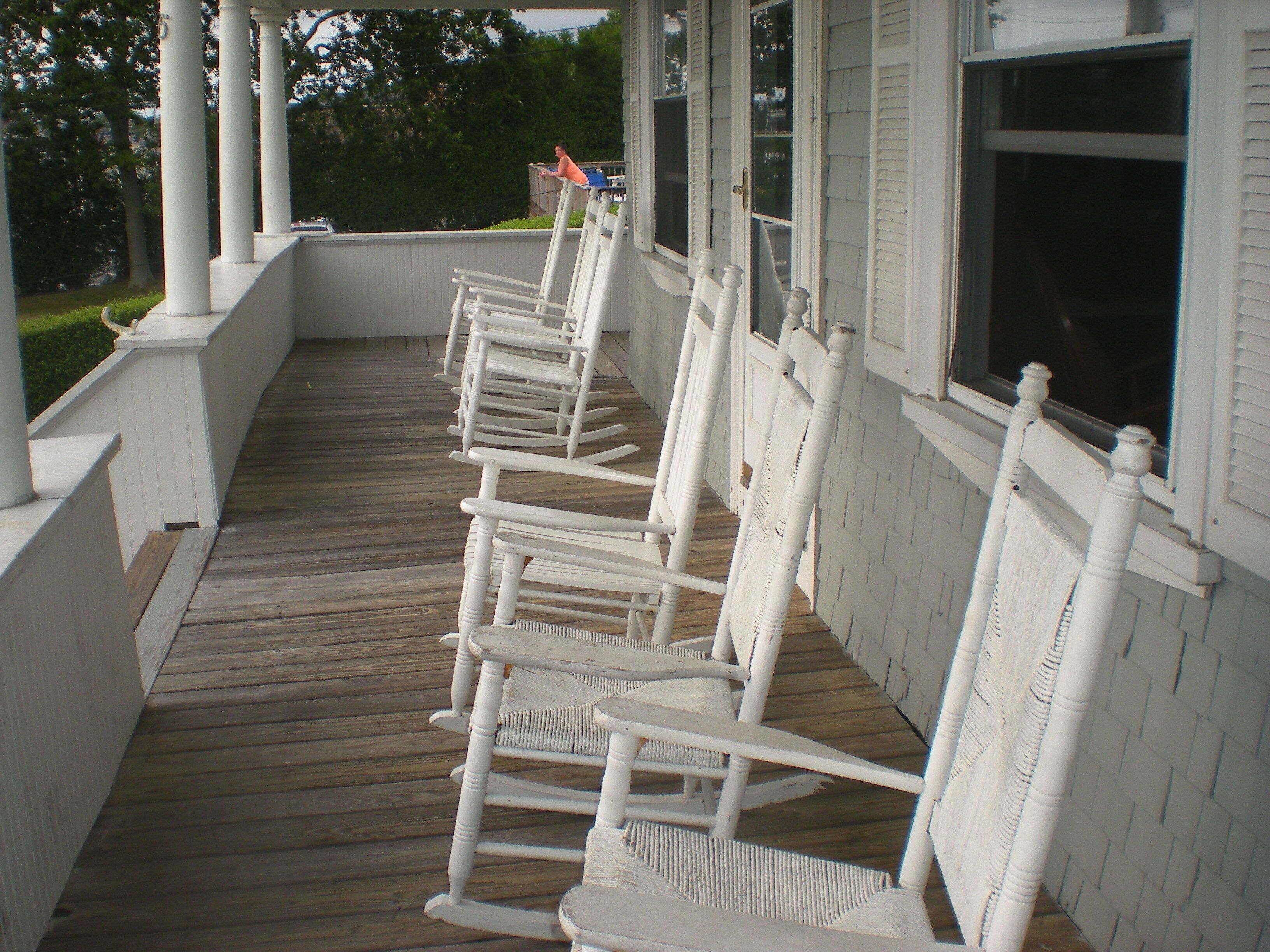 A covered porch with white wooden chairs and a wooden floor.