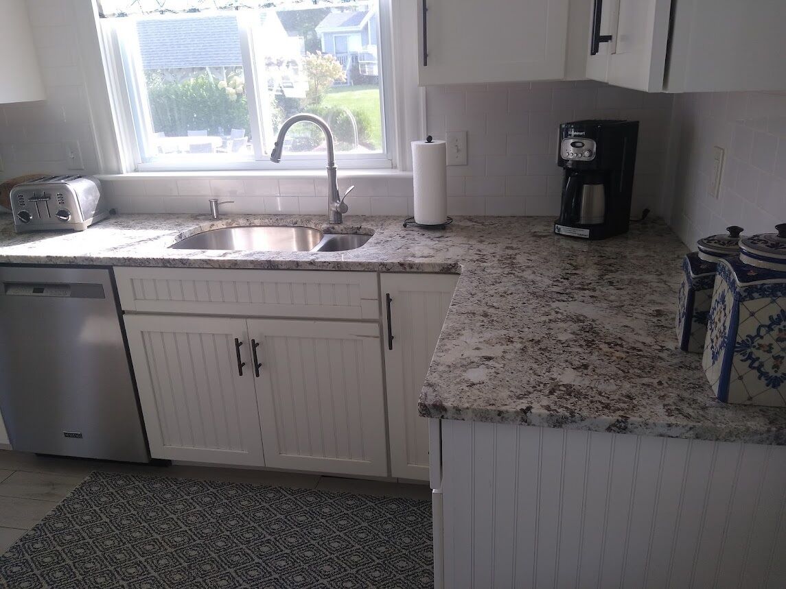 A kitchen with white cabinets, a granite countertop, a toaster, a coffee maker, and a window with a view of houses.