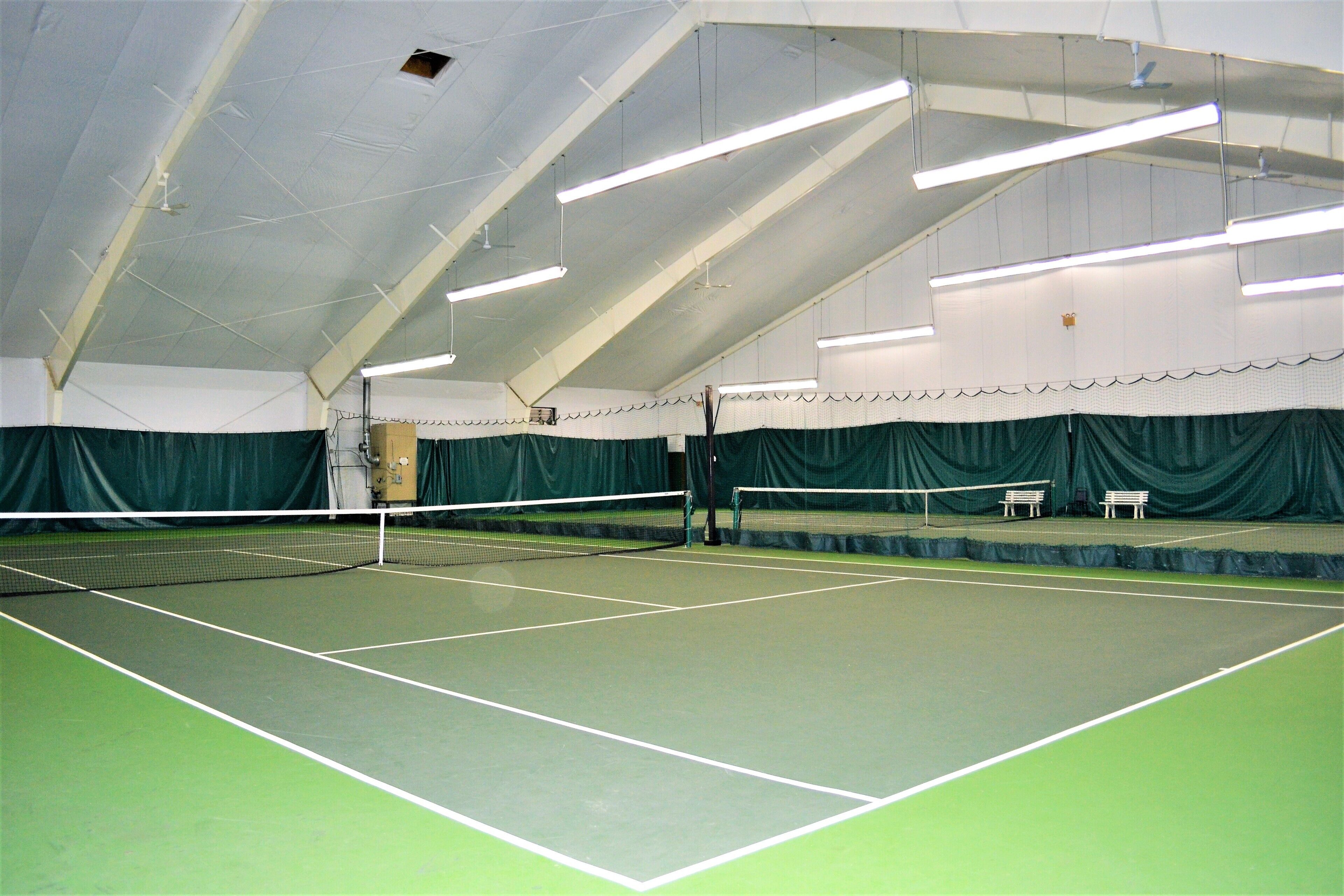 An indoor tennis court with a net, green flooring, and a high ceiling with lighting fixtures.