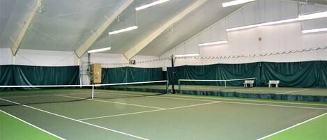 An indoor tennis court with a net, green flooring, and a high ceiling with lighting fixtures.