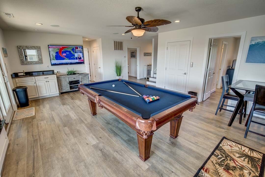 A pool table in a well-lit room with a ceiling fan, a television displaying a map, and a dining area with chairs and a table.