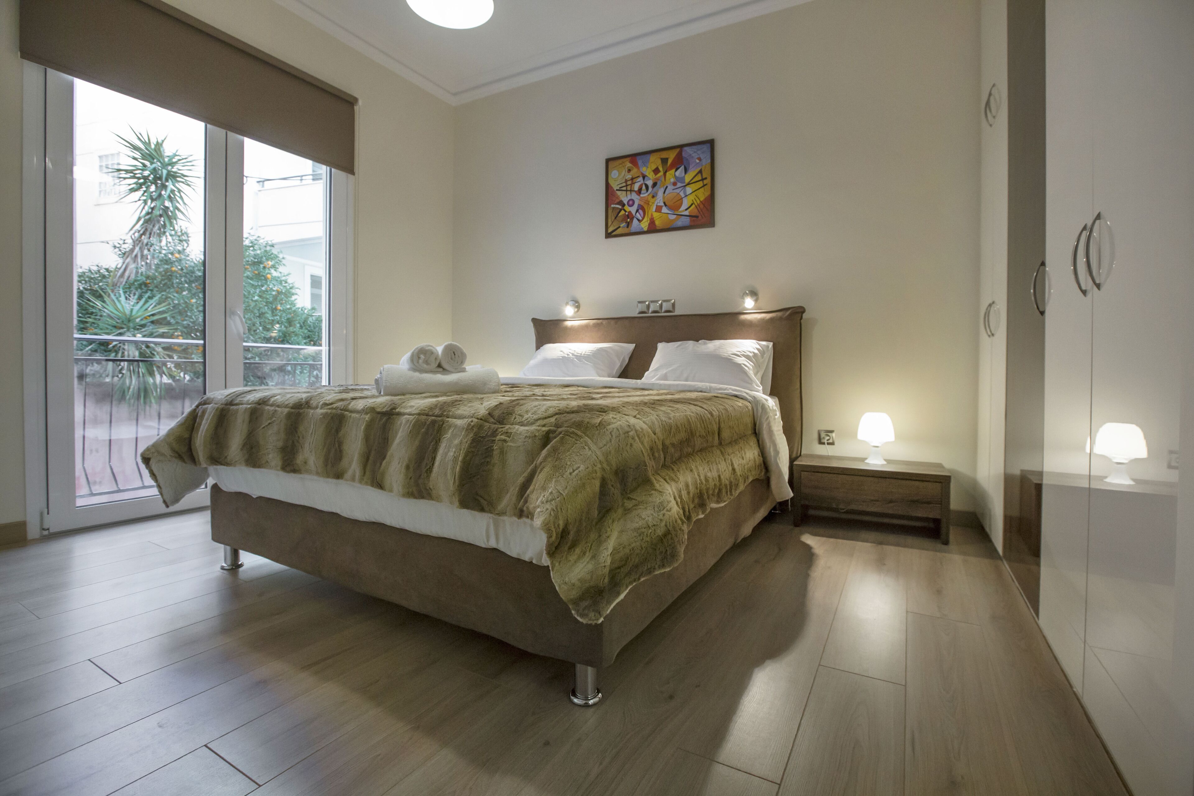 Ein Schlafzimmer mit einem großen Bett, einem Nachttisch mit Lampe, einem Gemälde an der Wand und Blick ins Freie durch eine Schiebetür.