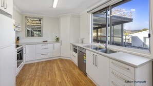 A modern kitchen with white cabinetry, a wooden floor, a built-in oven, a dishwasher, and a large window with a view of a house and trees. - Room for the whole family (Eildon)