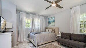 Un dormitorio con cama, un sofá gris, ventilador de techo y ventana con cortinas.