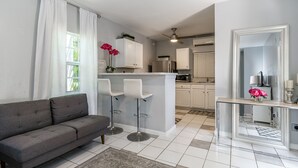 Un salón moderno con un sofá gris, dos taburetes blancos, una mesa de centro de vidrio y una cocina con armarios blancos y un refrigerador de acero inoxidable.