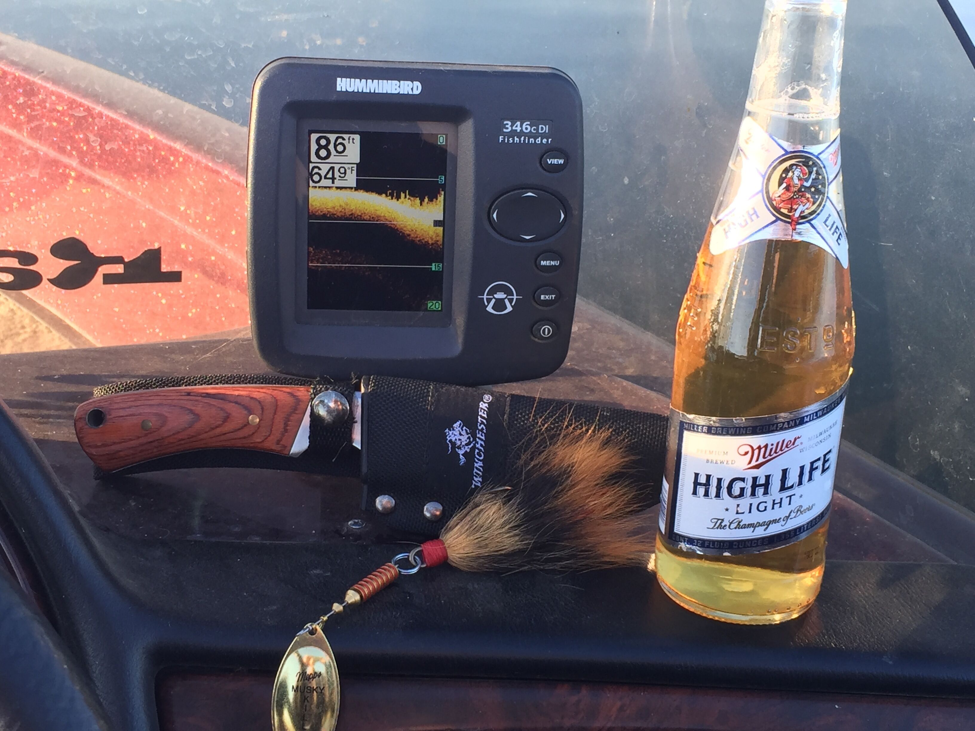A fish finder device, a bottle of Miller High Life beer, and a fishing lure on a vehicle dashboard.