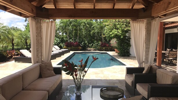 A covered outdoor area with a pool, lounge chairs, and a table with a vase of flowers.