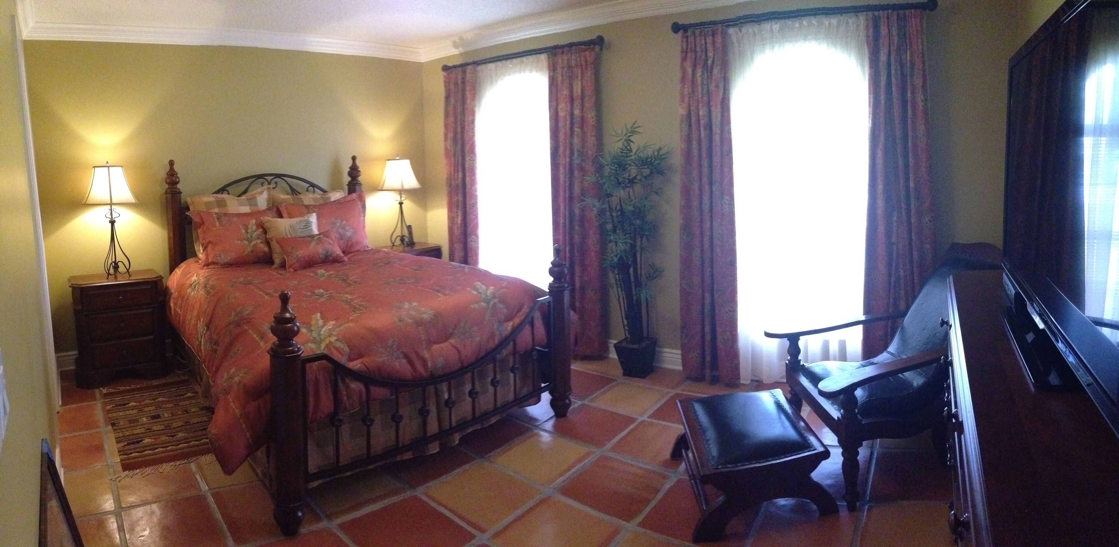A bedroom with a bed, two bedside lamps, a potted plant, a piano, and a window with curtains.