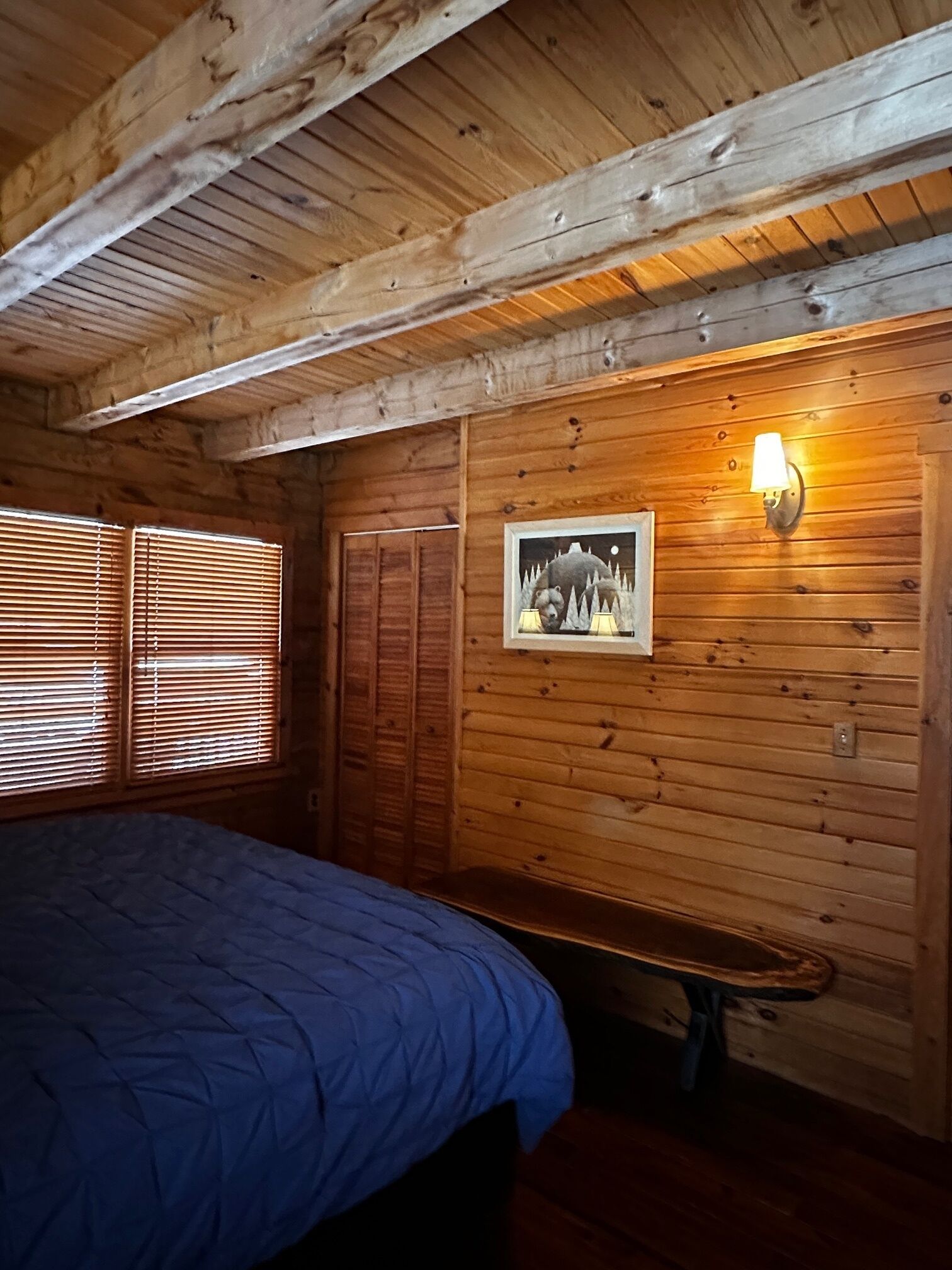 Une pièce de cabane en bois comprenant un lit, un banc, une lampe fixée au mur et une image représentant un ours et des arbres.