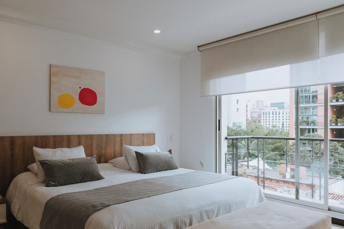 A modern hotel room with a large bed, a wooden headboard, a bedside table with a phone, and a view of the city through large windows.
