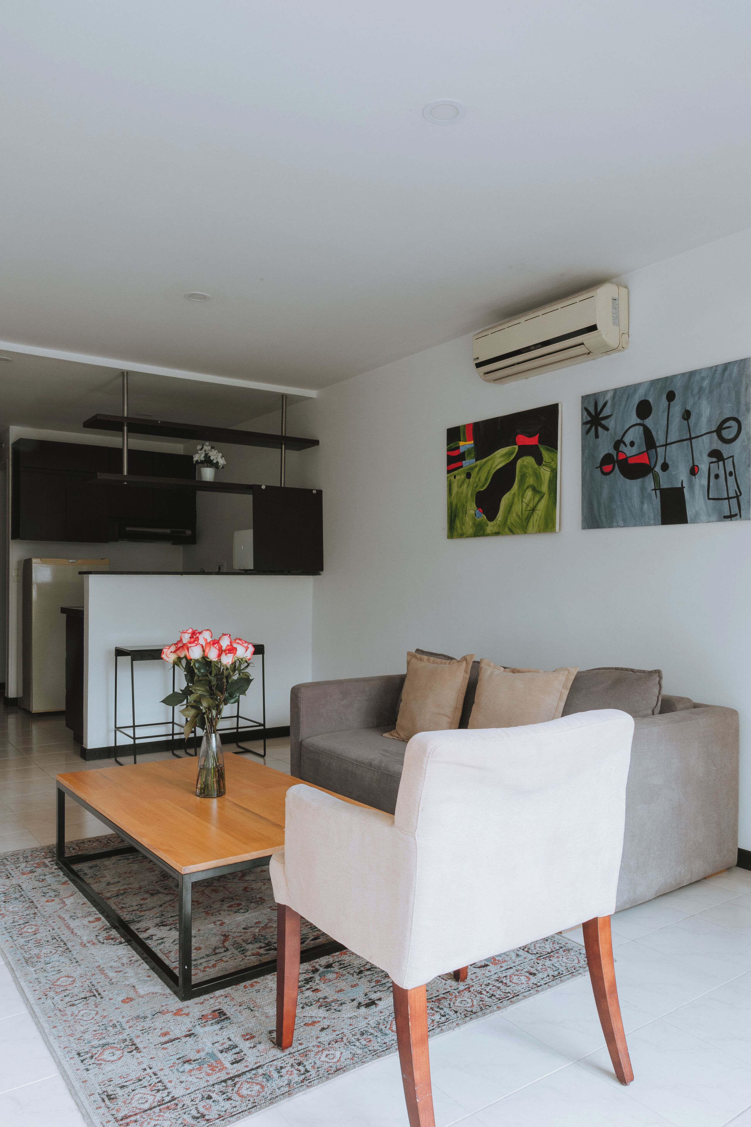 Een moderne woonkamer met een bank, een salontafel en een bloemenvaas. Er is een keukenhoek met een televisie en een flatscreen. De muren zijn versierd met abstracte kunst.