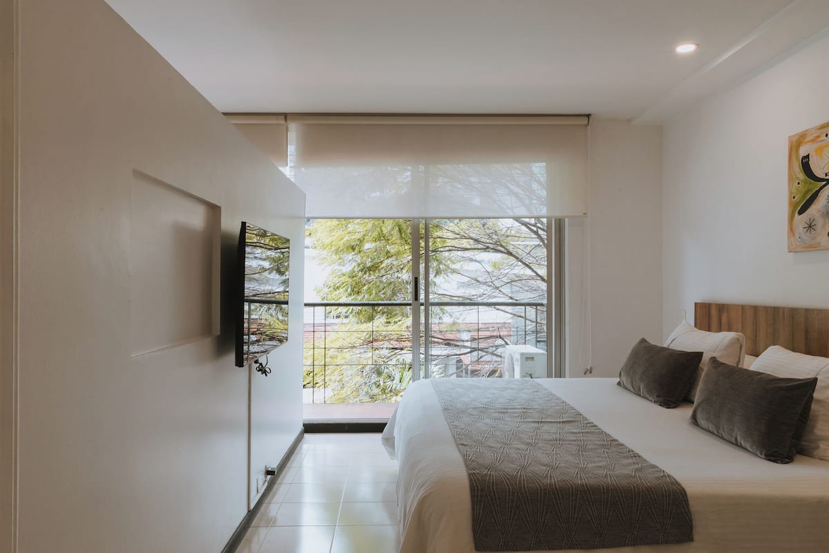 A modern bedroom with a large bed, a flat-screen TV mounted on the wall, and a window offering a view of trees and buildings.
