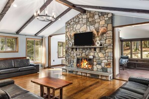 A spacious living room with a stone fireplace, leather furniture, and large windows offering a view of the outdoors.