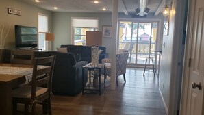 A living room with a TV, a dining table, and a sofa. There is a balcony with a railing and a view of the outside. -  Penthouse Suite in the heart Sylvan Beach Resort (Sylvan Beach)