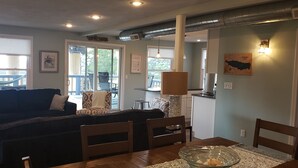 A living room with a dark-colored sofa, wooden chairs, a glass coffee table, and a dining table. There is a sliding glass door leading to a balcony with outdoor furniture. -  Penthouse Suite in the heart Sylvan Beach Resort (Sylvan Beach)