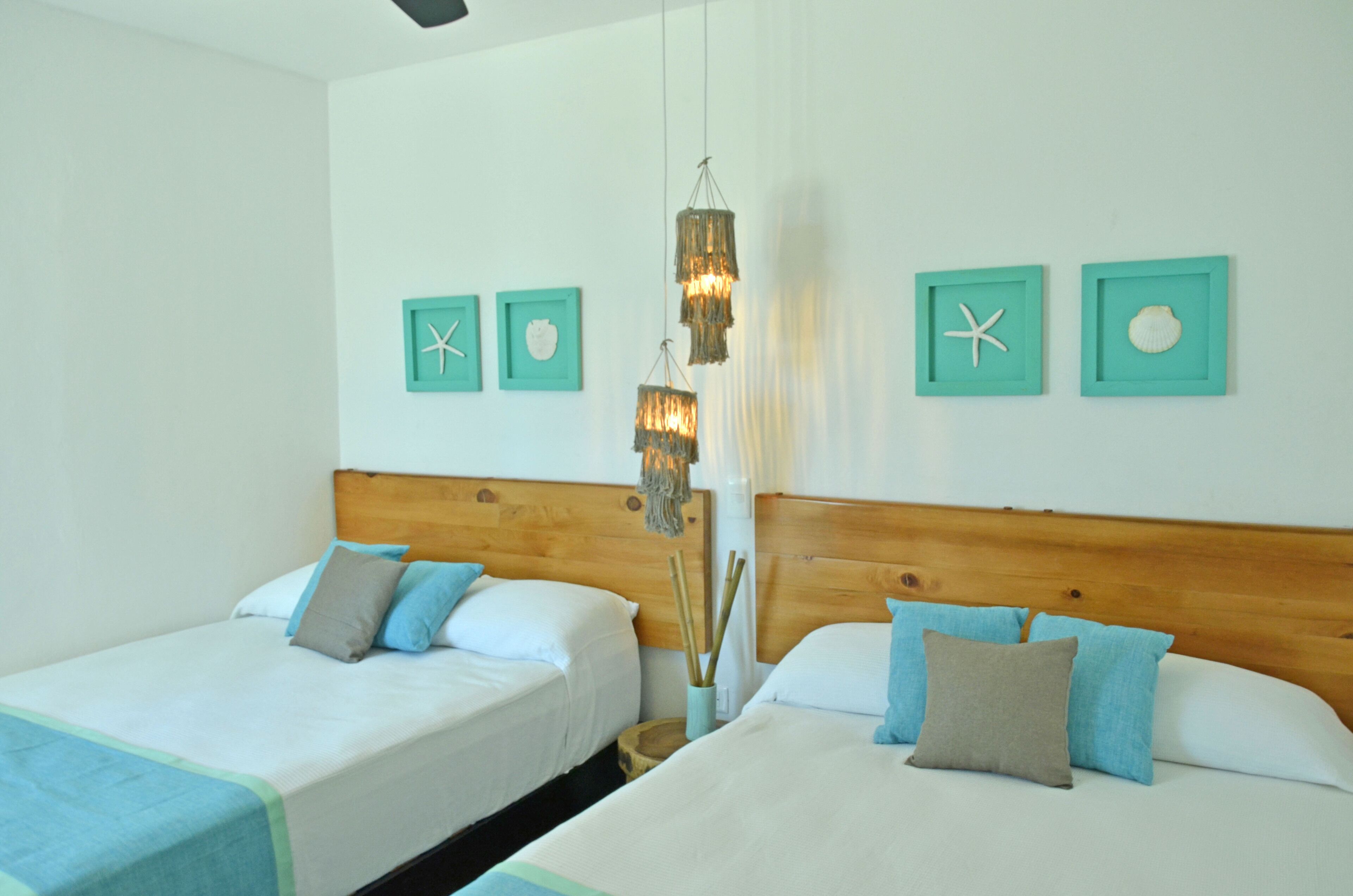 Une chambre d’hôtel avec deux lits, une tête de lit en bois, des décorations murales encadrées de motifs turquoise et un luminaire suspendu.