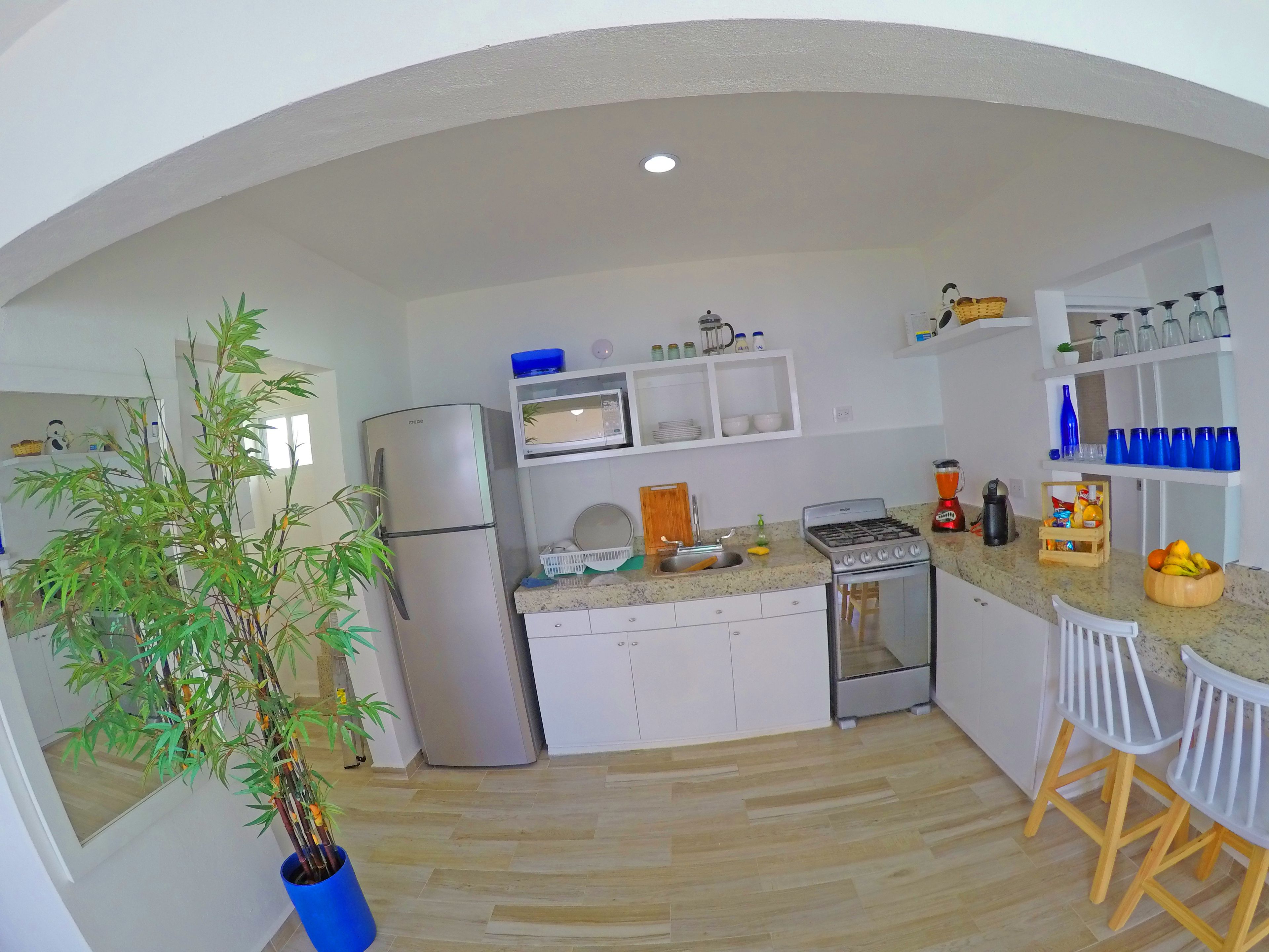A modern kitchen with white cabinets, a stainless steel refrigerator, a microwave, a gas stove, and a sink. There are two white bar stools and a potted plant.