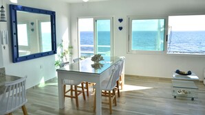 Un comedor con una mesa de vidrio, sillas blancas, un ventilador de techo y vistas al océano a través de amplios ventanales.
